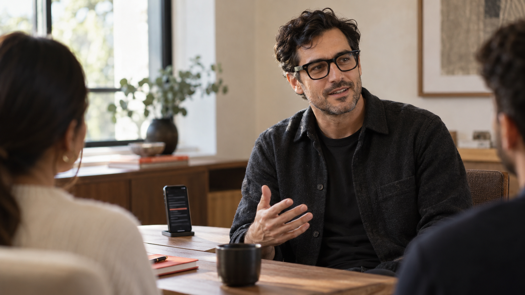 AI-generated editorial image of a speaker wearing black smart glasses in a small meeting, with an iPhone prompter nearby